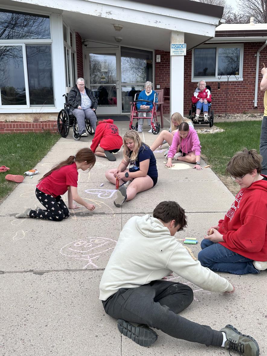 Residents (from left) Sonja Goembel, Marilyn DeBates and Anna Harrison enjoy watching the visiting sixth-graders create sidewalk art with colorful chalk. Students are (from foreground to back) Jack Bailey, Levi Baker, Harper Hulstein, DesiRae Miller, Lucy Knips, Avery Van’t Hof and Sydney Van Grootheest. Lori Sorenson/Rock County Star Herald Photo